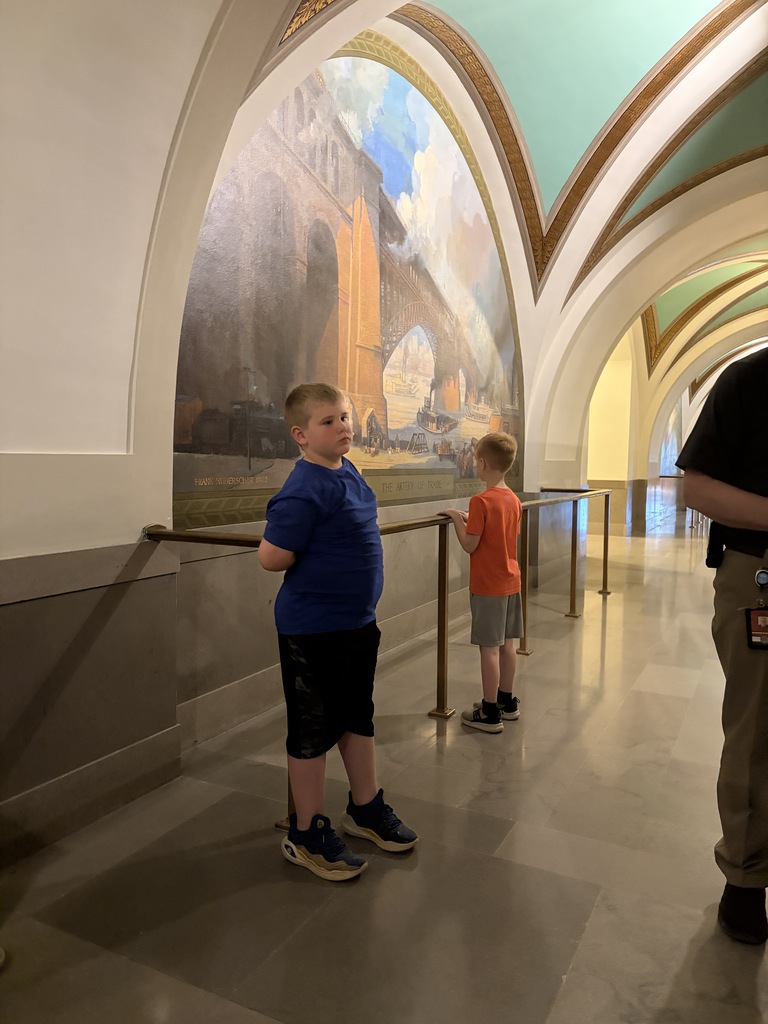 Two children, one wearing a blue shirt and the other orange, stand in a hallway with a large mural behind them.