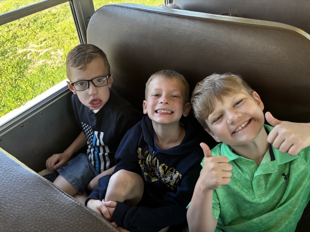 Three boys sitting in a school bus with one boy giving a thumbs up and two smiling.