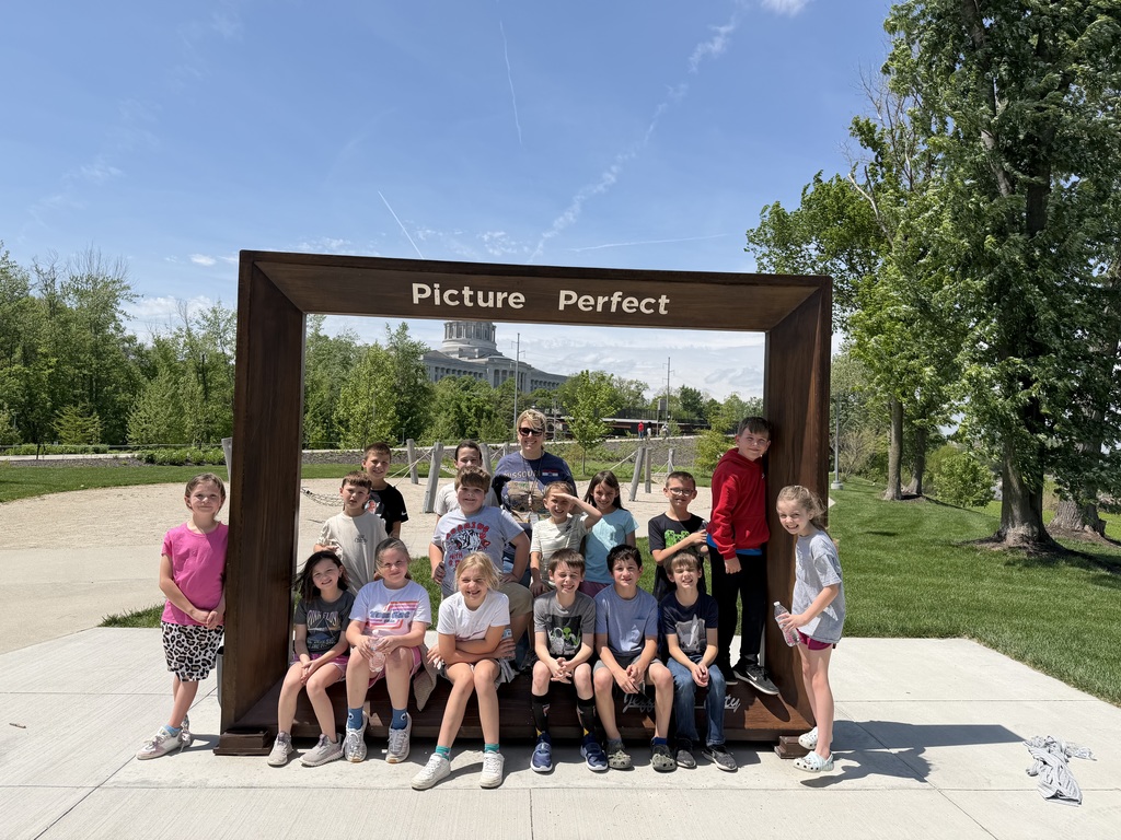 Kids and two adults pose for a photo in a photo frame, with a grassy area and trees in the background.