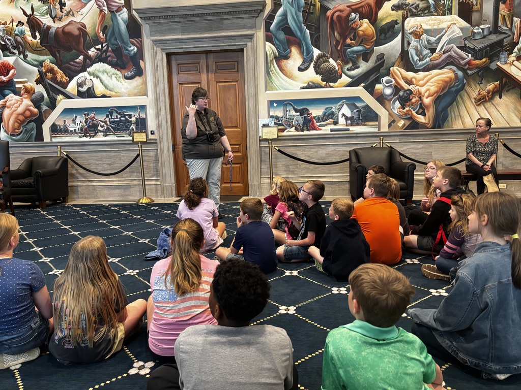 A group of children listening to a woman speak, in a large room with patterned flooring and large murals.
