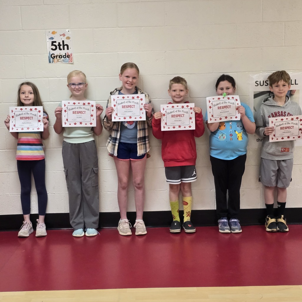 Seven kids standing in a row hold certificates, smiling for a photo. A red floor and white wall in the background.