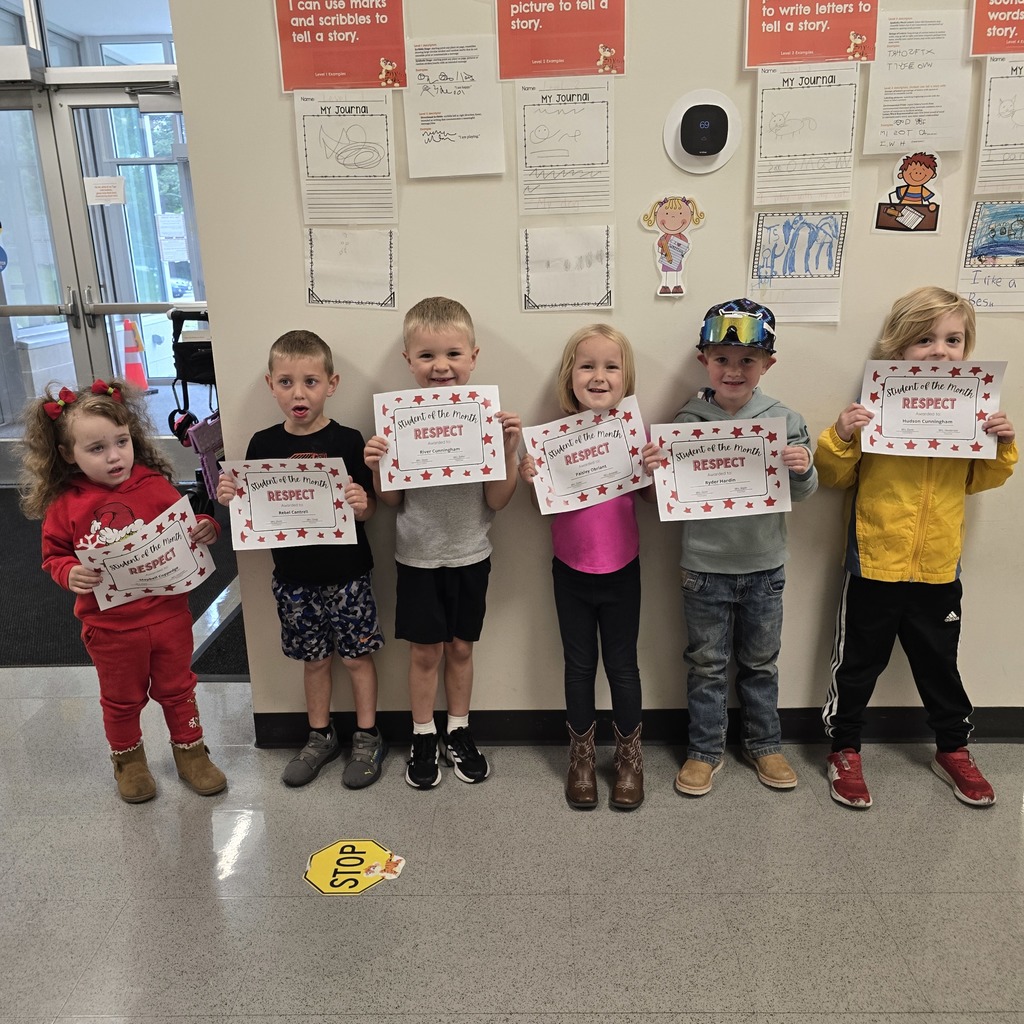 Seven children stand in a line, smiling and holding certificates, in front of a wall with papers and a stop sign on the floor.