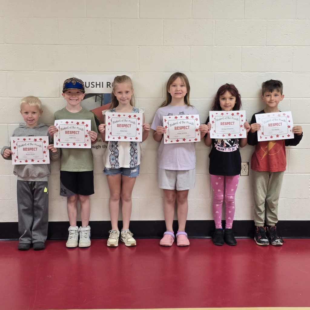Group of children holding certificates, standing in a row on a red floor, next to a white wall.