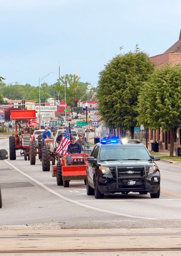 Tractors and a police car with flashing lights drive down a street with buildings, trees, and a clear sky.