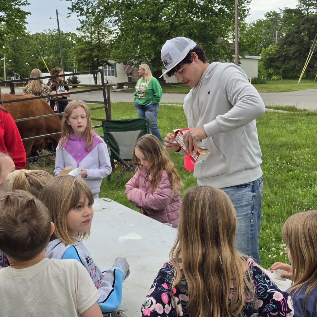 Children sit around a table while a man in a white cap and gray hoodie hands out items.