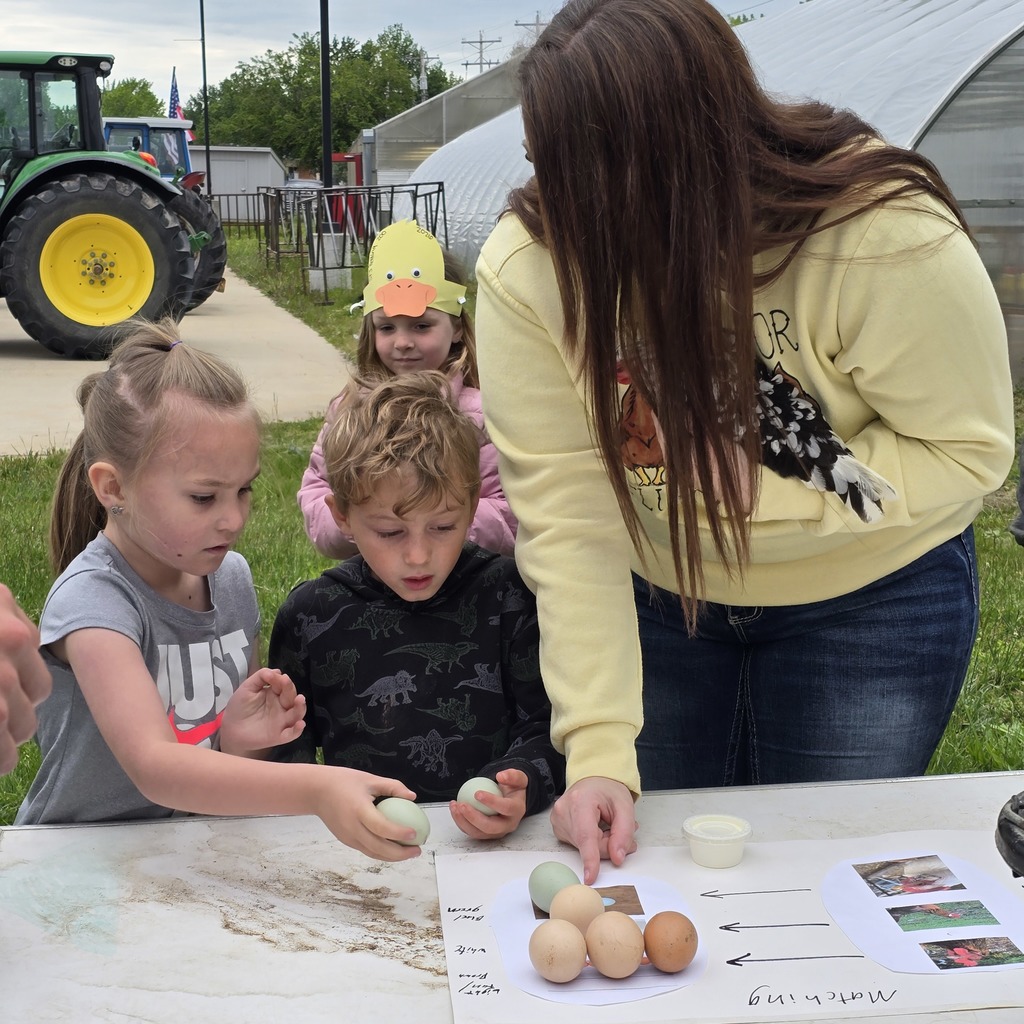 Adult and children at a table with eggs, near a tractor. One child wears a rubber duck hat.