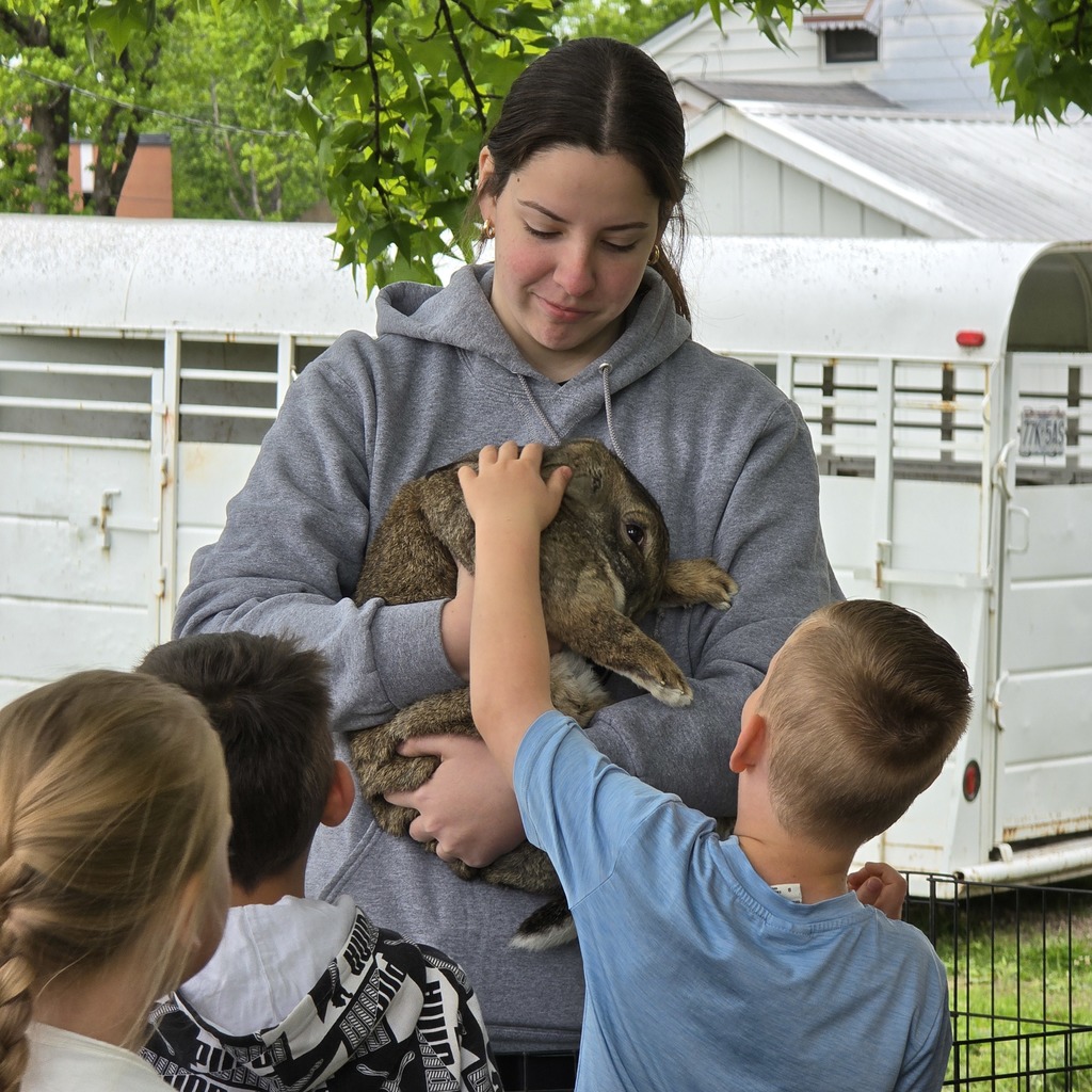 A woman in a gray hoodie holds a rabbit while four children gather around. A trailer is in the background.