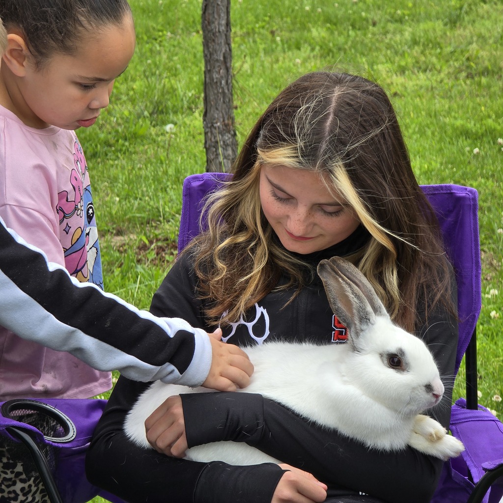 Two young girls hold a white rabbit; one seated in a purple chair, the other standing, both in a grassy area.