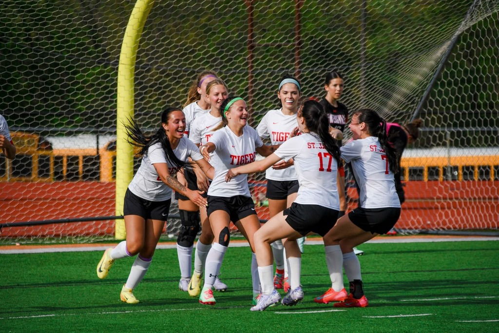A group of young women in white uniforms, celebrating on a field with a soccer goal in the background.