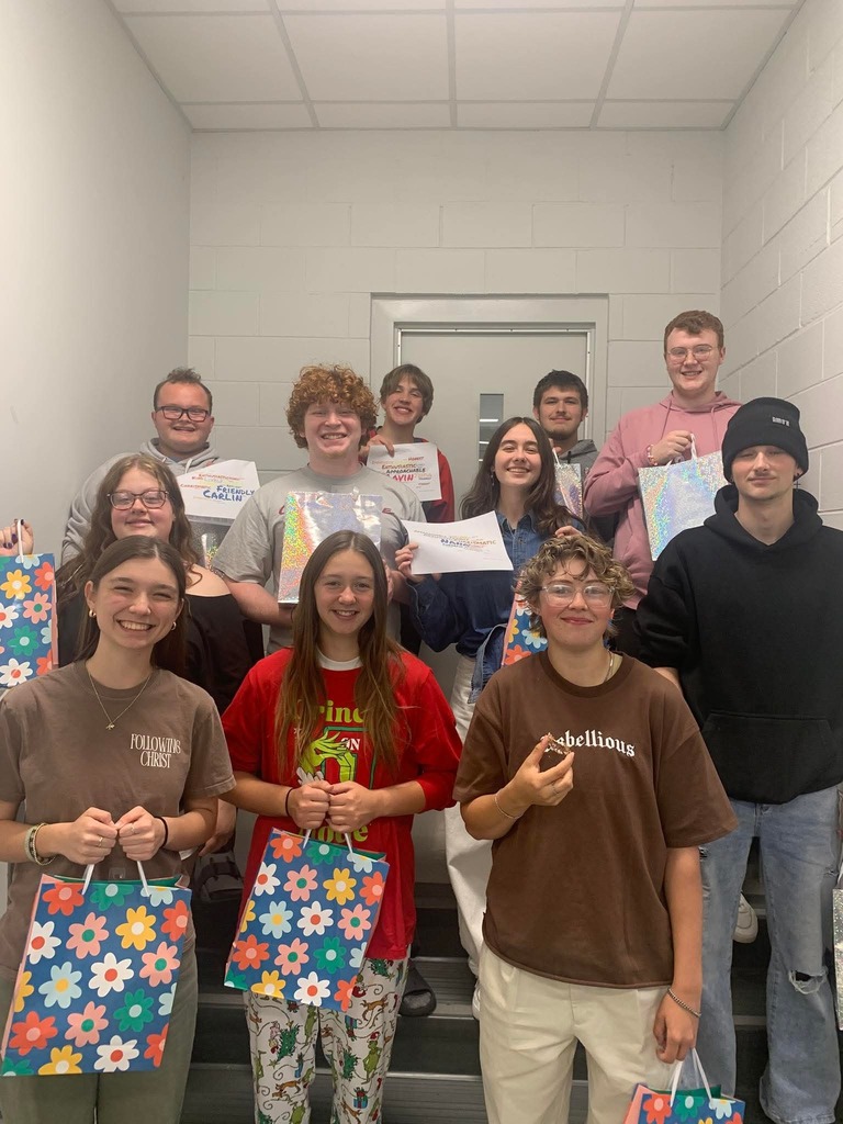A group of young adults with various expressions hold up bags and certificates in a hallway with white walls and a door.