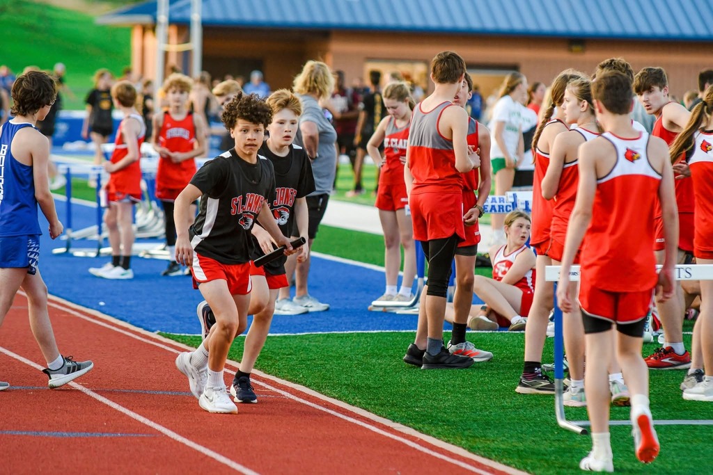Several children in red uniforms are running on a track with hurdles. Some are standing on the sidelines.