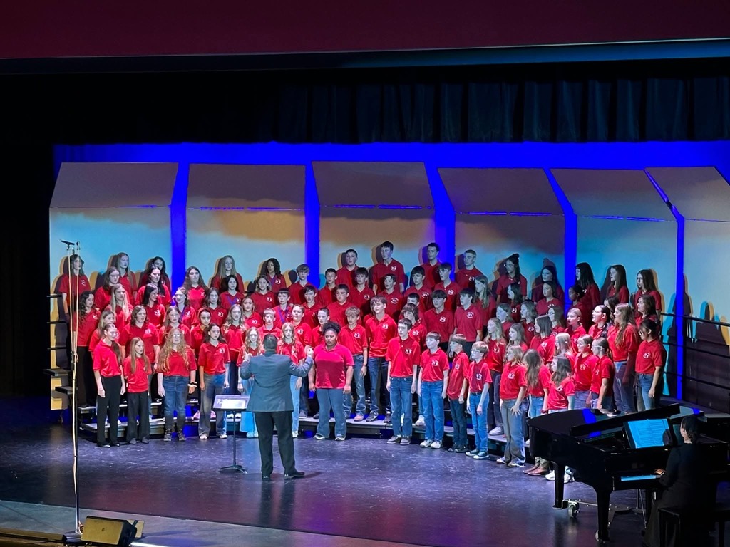 A large group of people in red shirts stand on stage with a piano and blue lighting.