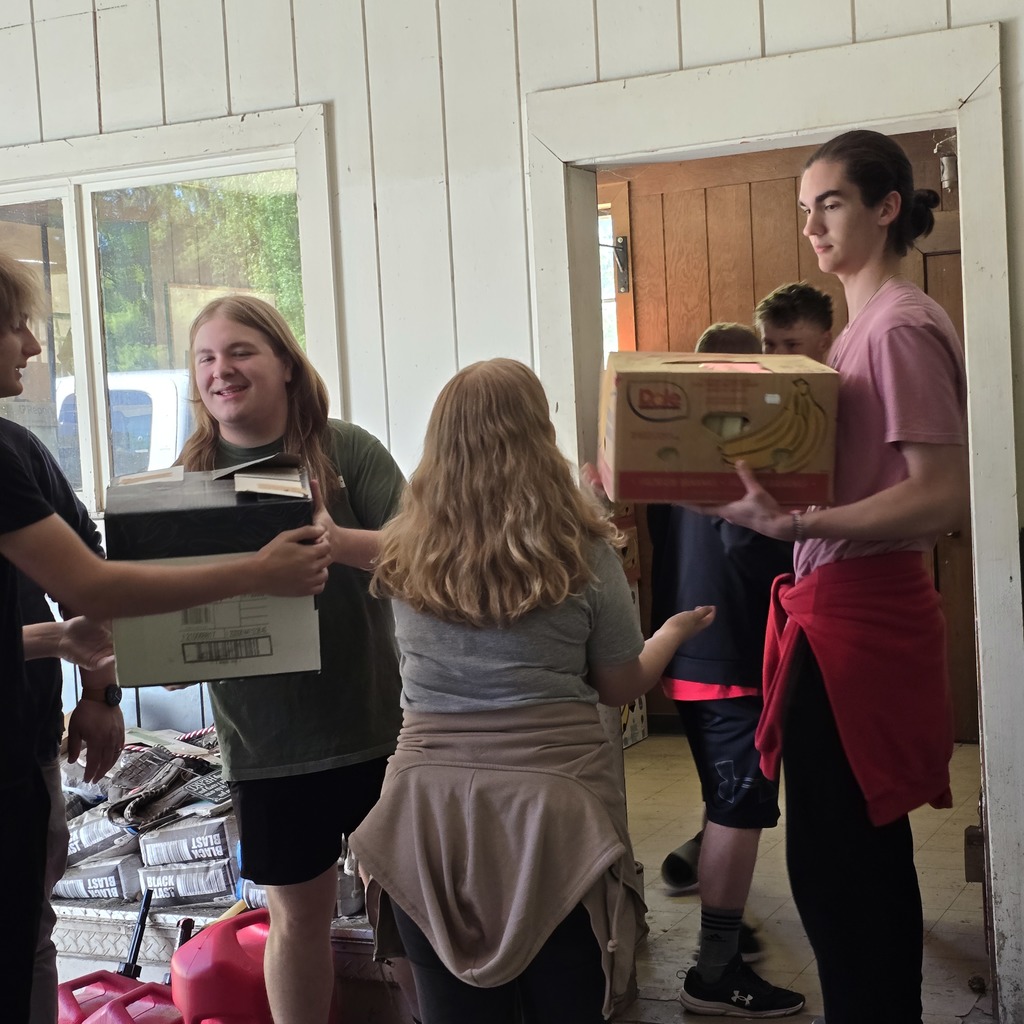 Five people in a kitchen. Three carry boxes, one holds a box of bananas. Piles of boxes behind them.