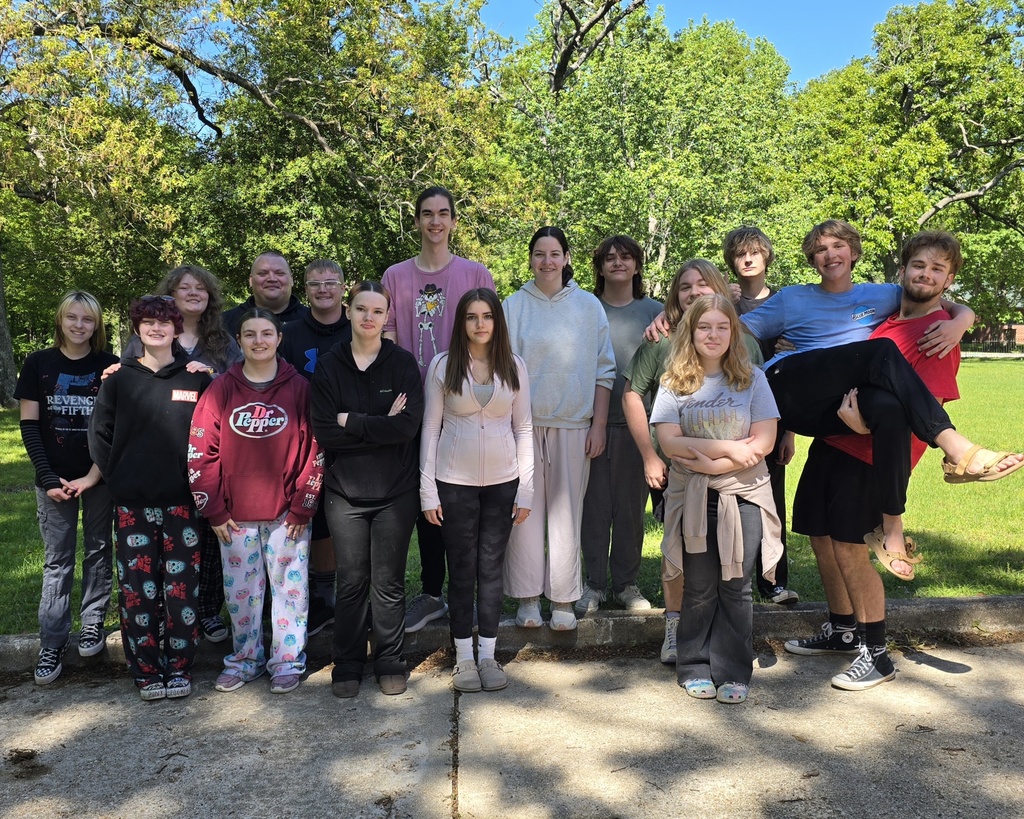 A group of young people stands together on concrete, wearing various clothing. Green trees and grass are in the background.