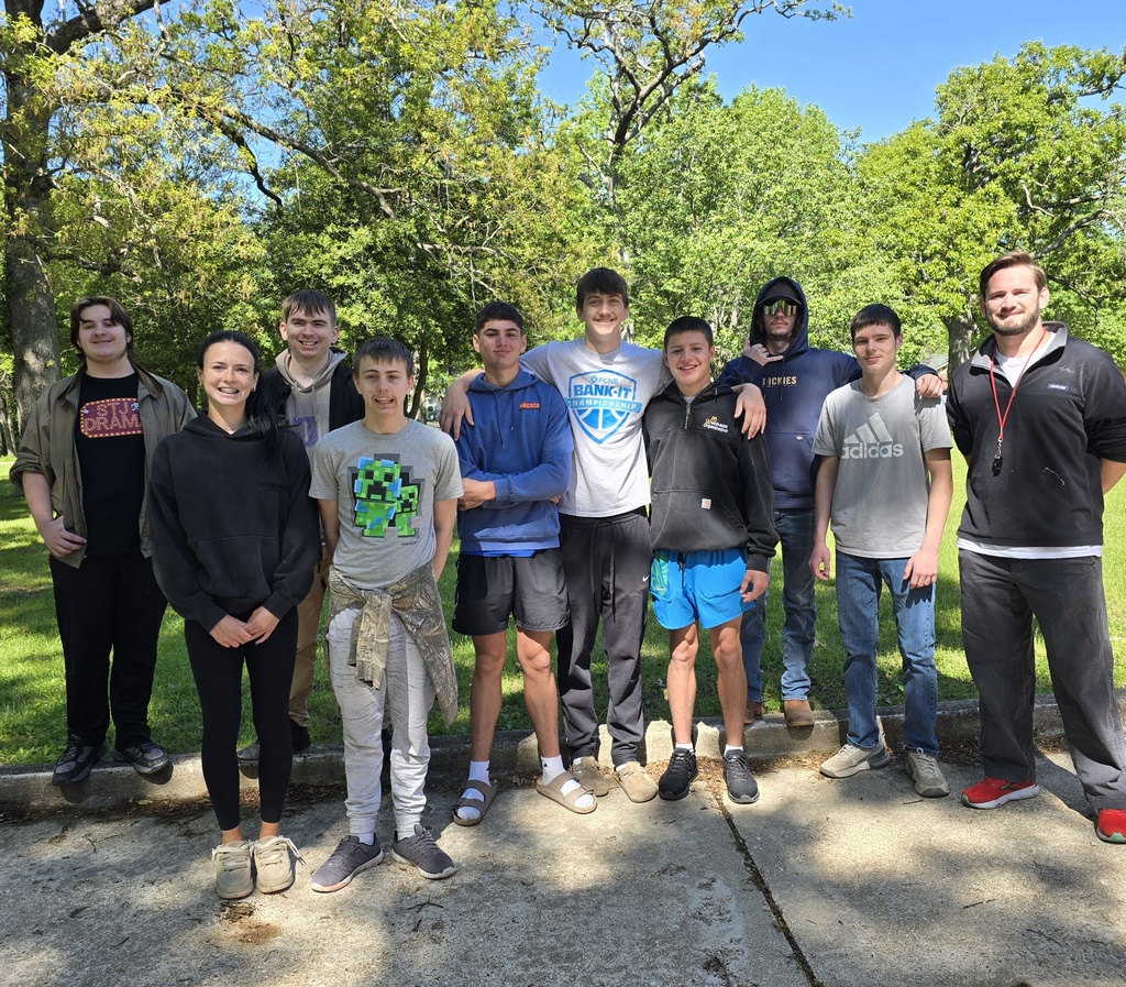A group of young people stands together on concrete, wearing various clothing. Green trees and grass are in the background.