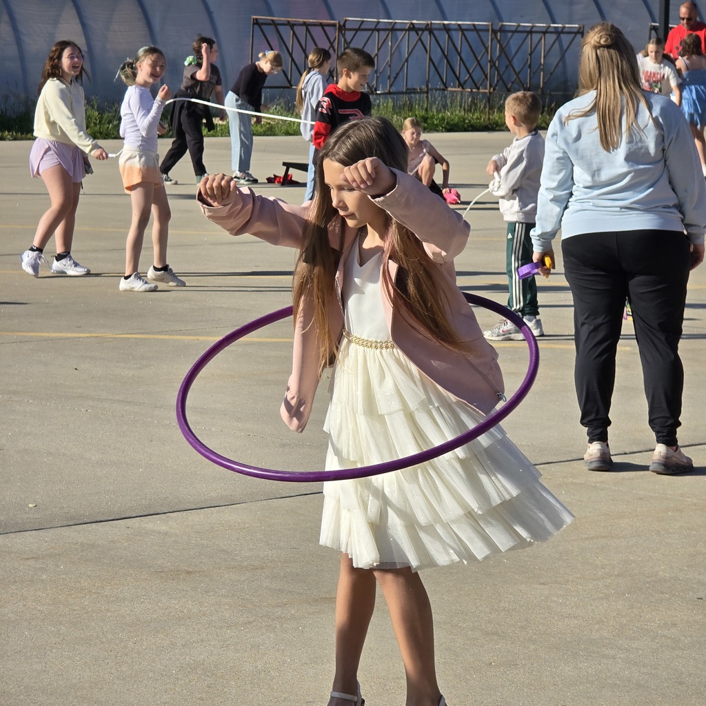 A young girl in a white dress and purple hula hoop spins it while others walk behind.
