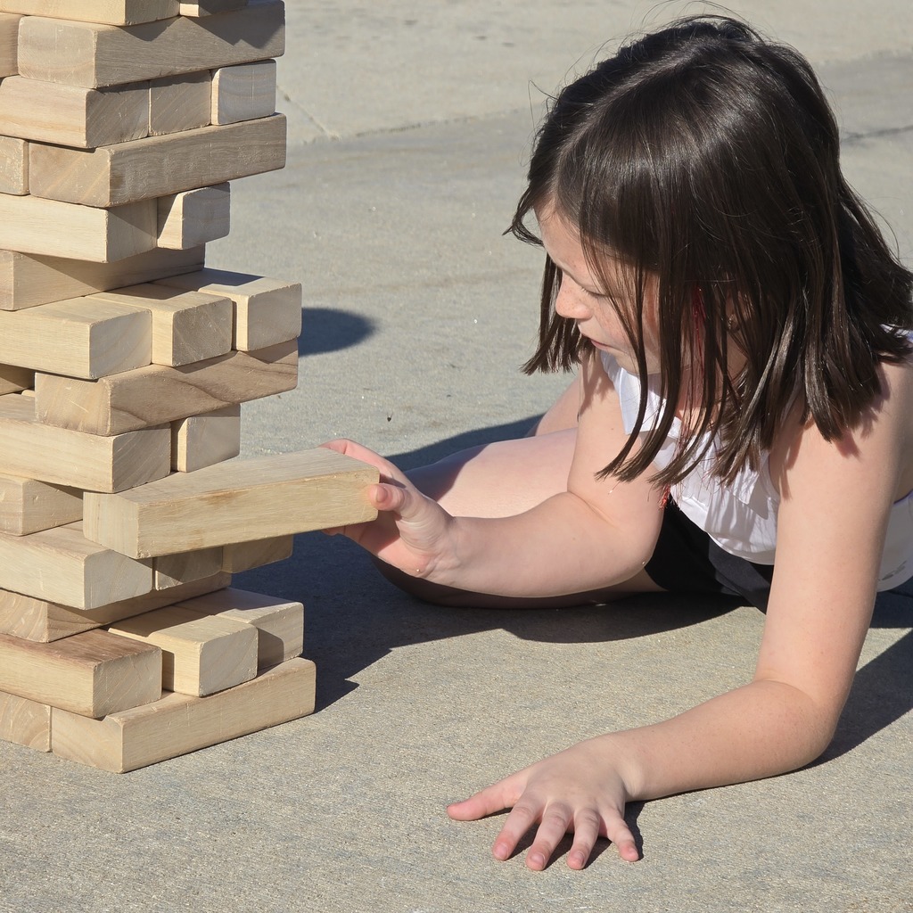 A child is lying on concrete, attempting to remove a block from a tall wooden tower.