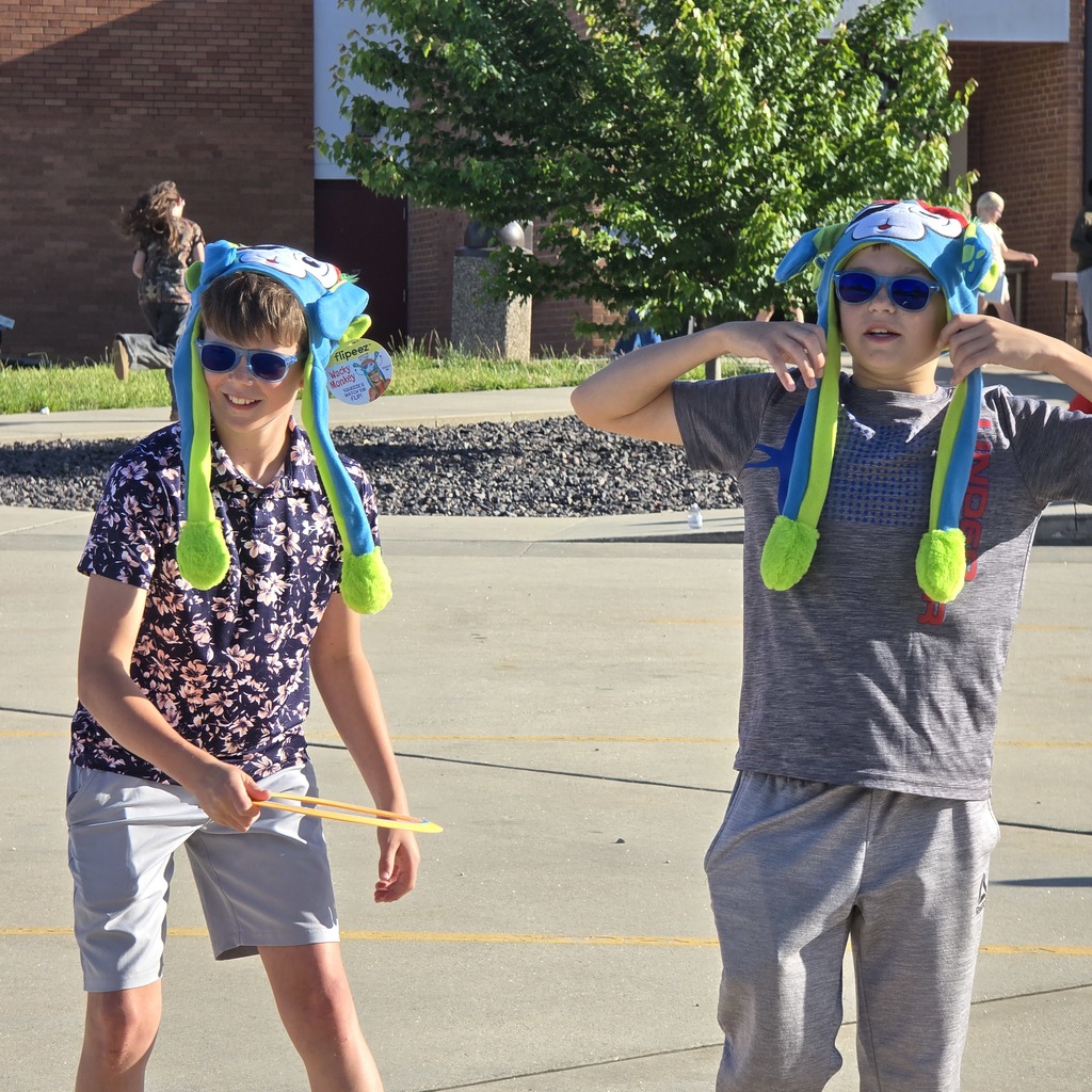 Two boys wear sunglasses and animal ear hats, one holds drumsticks. Behind them is a building and a tree.