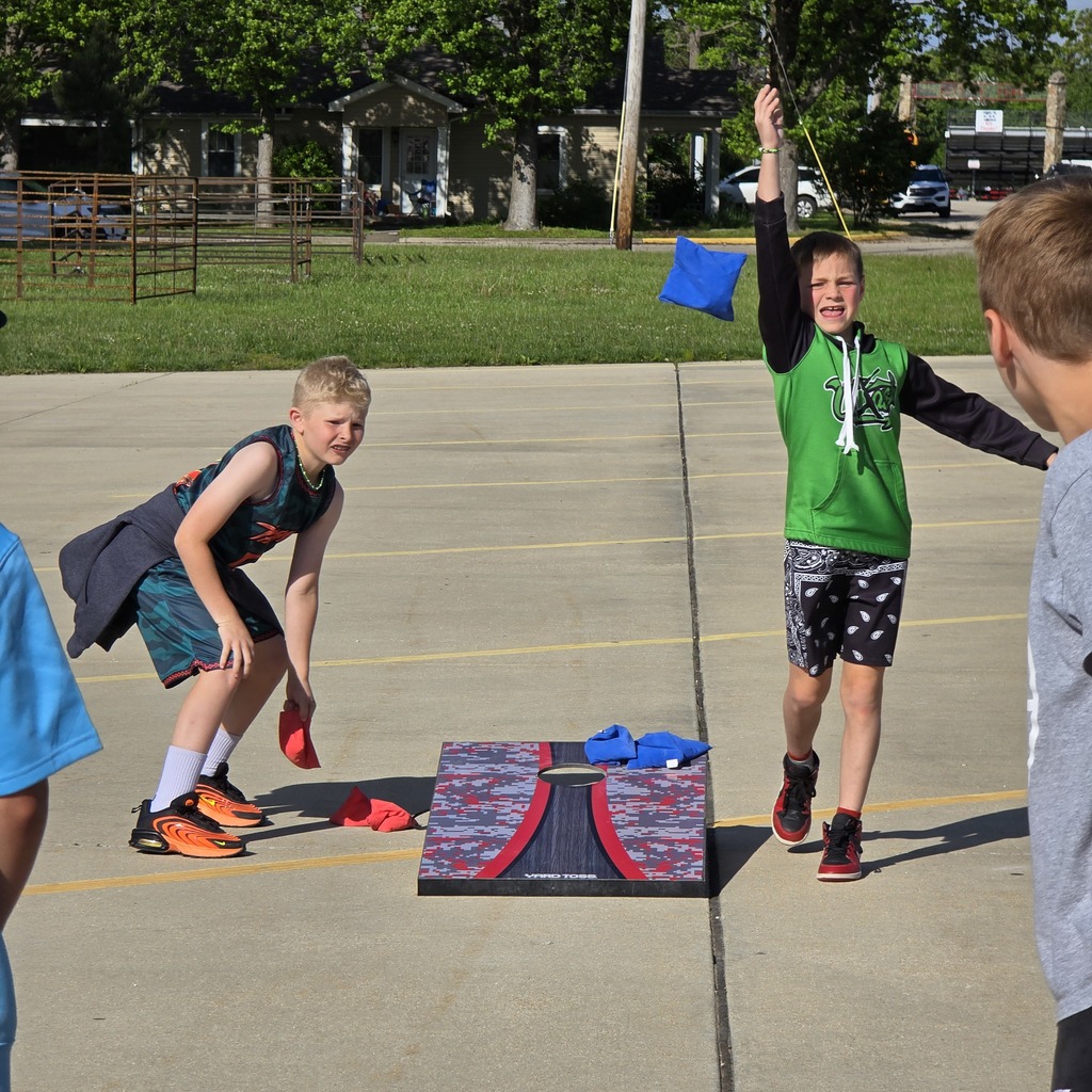 Three boys play cornhole on a paved area with a grassy background. One boy prepares to throw.