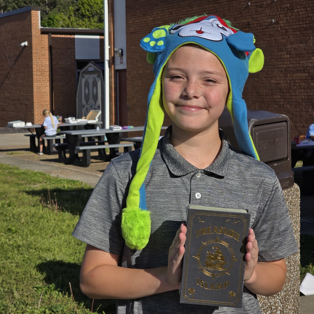 Child in hat and gray polo holds book with golden design. Behind, picnic tables, brick building, and a person.