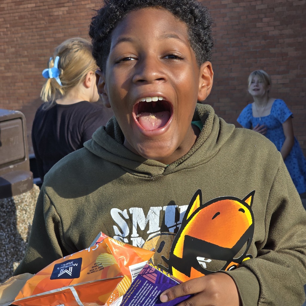 A young person in a green hoodie holds a bag of snacks while laughing, with two women in the background.