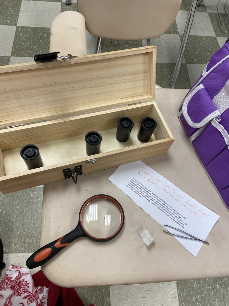 Magnifying glass, small black lenses, and paper are on a table with a purple bag. Checked floor in background.