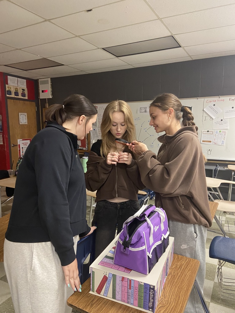 Three students examine a phone held by one of them, standing near a table with a bag and books.