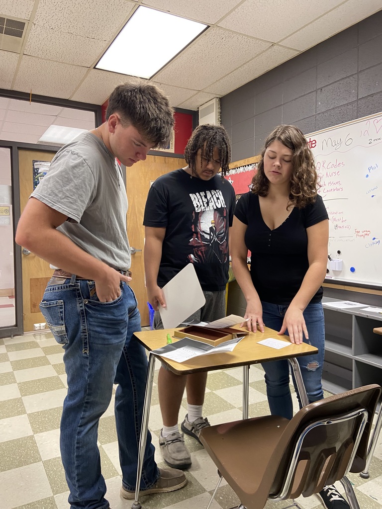 Three people standing around a desk in a classroom, looking at papers, with a whiteboard behind them.