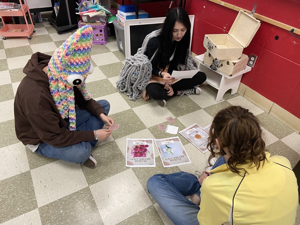 Three people sit on a floor with a white and black checkerboard pattern. Two have headwear.