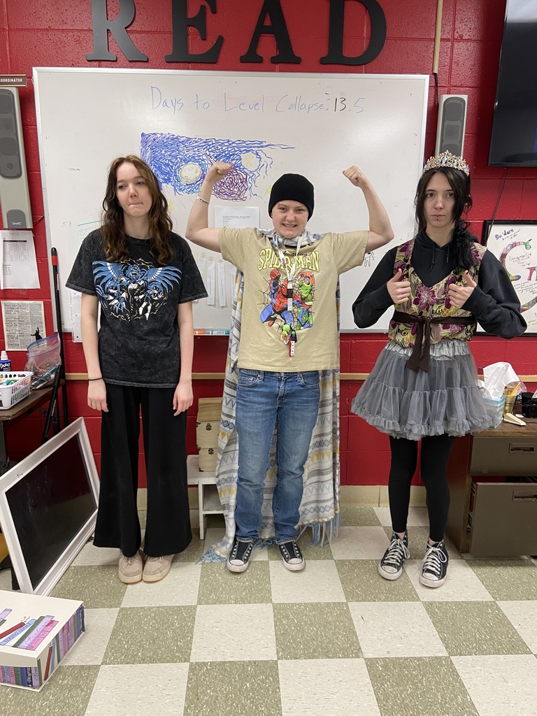 Three students in a classroom with one raising both arms. The word "READ" is on the wall behind them.