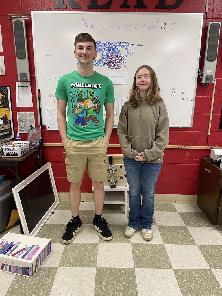 Two students in a classroom with a whiteboard labeled "READ" and a checkered floor.