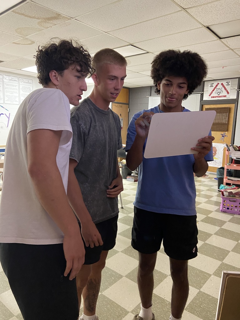 Three male students stand in a classroom examining a paper.