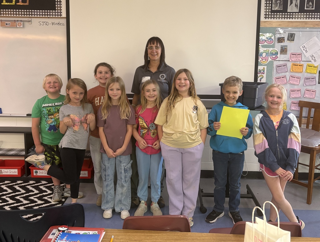A group of young students in a classroom, standing and smiling with a teacher. The background includes a whiteboard and bulletin board.