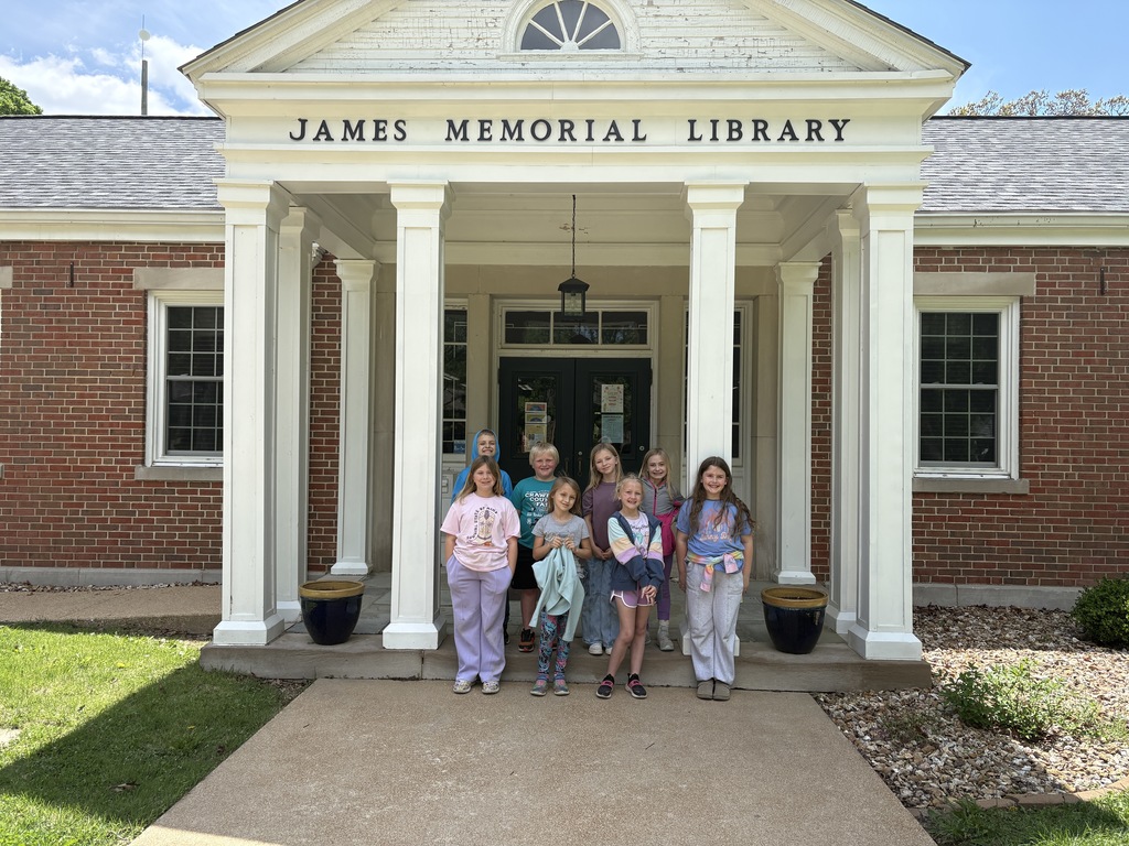 A group of children stand on the porch of a brick building with white columns. The sign reads "James Memorial Library."