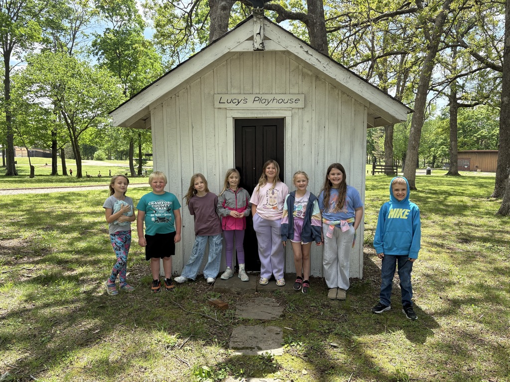 Several children stand in front of a small white building with trees in the background.