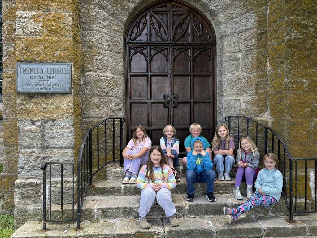 A group of children sitting on steps in front of a church door, with a stone wall and sign.
