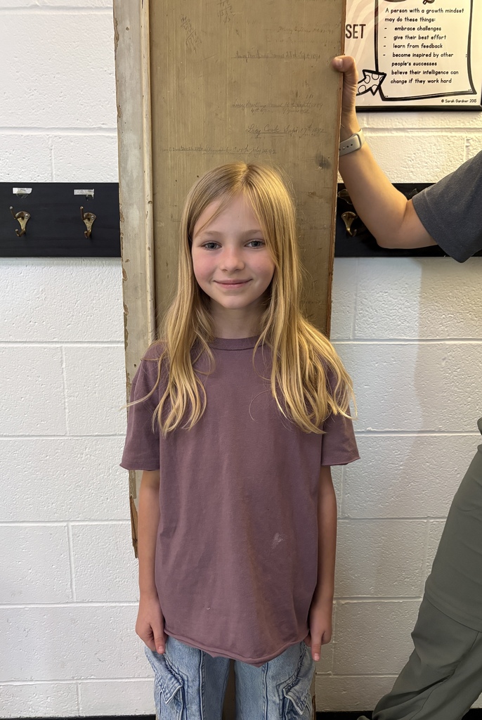 A young girl with blonde hair smiles, standing next to a door with a poster. She wears a purple t-shirt and blue jeans.