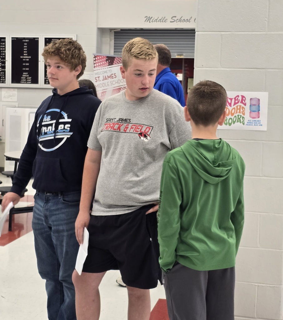 Three young males in school attire, standing and conversing. One holds a piece of paper. A pink sign reads "Never Give Up."