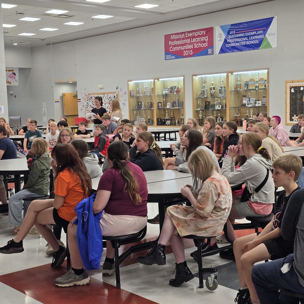 A classroom with students sitting around tables; some wear sneakers and one person has a backpack.