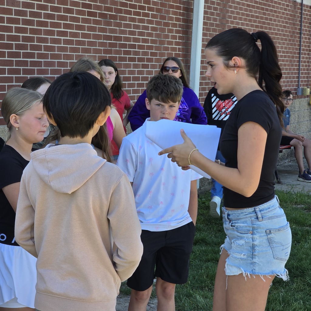 A group of students outdoors; one holds a paper and points. Brick building behind them.