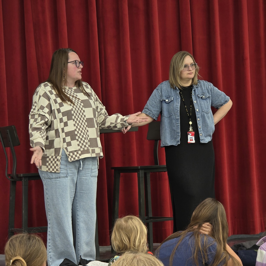 Two women stand facing each other, one wearing glasses and a patterned jacket, the other in a denim jacket.
