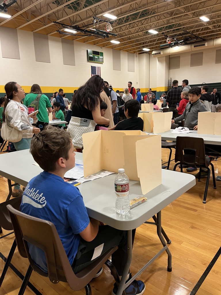 A gymnasium with tables and chairs, a boy sits at a table with a clear divider. People stand and sit around tables.
