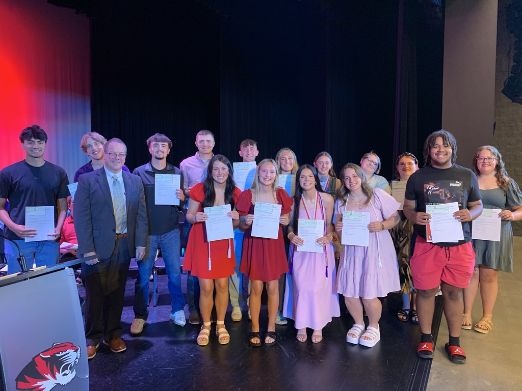 Group of students in front of a stage holding certificates, a podium on the left, and a logo.