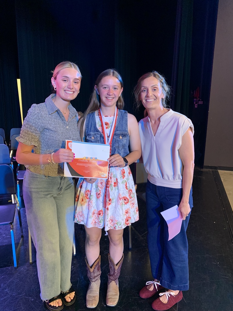Three women stand on a stage, one holding a certificate, another holding papers, and the third smiling. A black curtain is behind them.