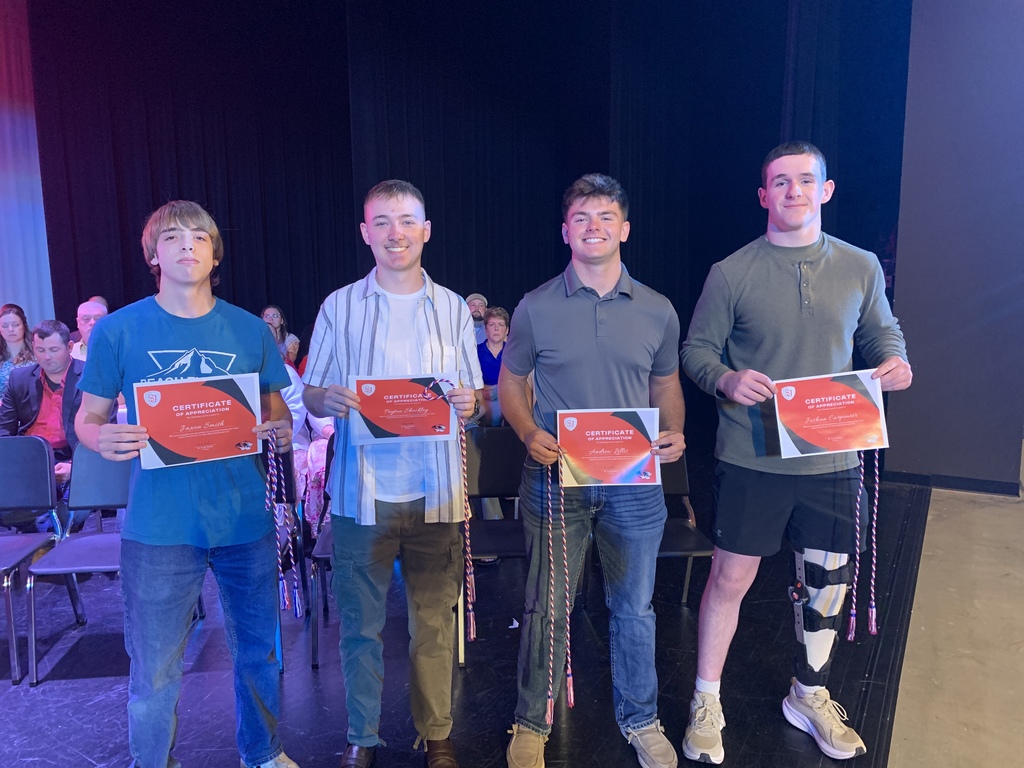 Four men hold certificates and stand together on a stage, with chairs and a crowd in the background.
