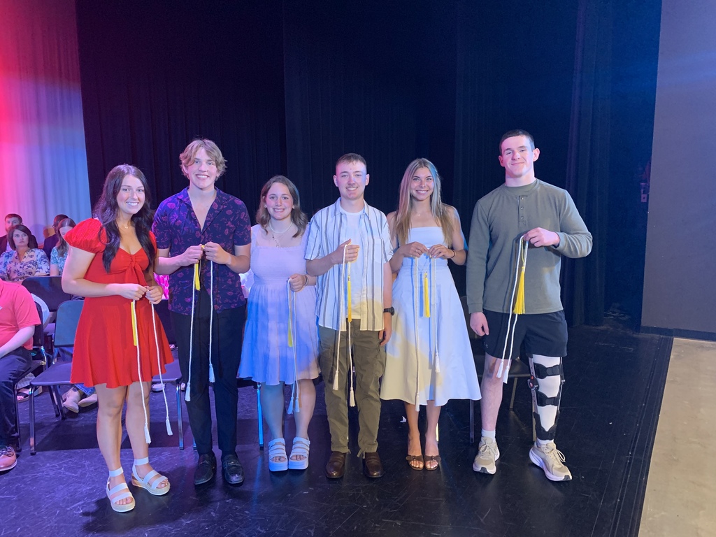 Six performers stand on a stage, holding yellow ribbons, with an audience seated in the background.