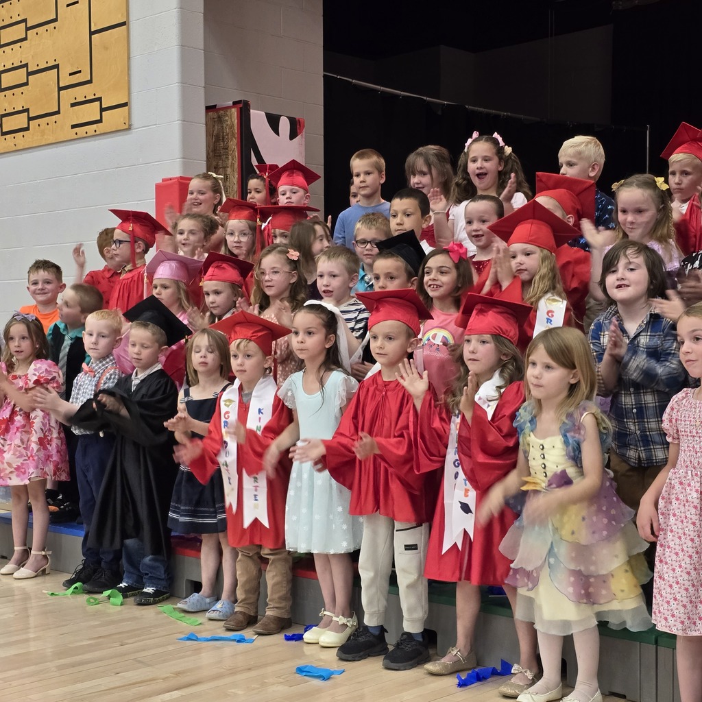 A group of children in red graduation gowns and hats stand on stage with a wooden floor.