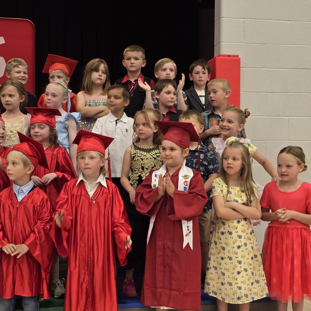 Children in graduation robes and dresses stand on stage. Some wear hats, others do not. Some wear sashes.