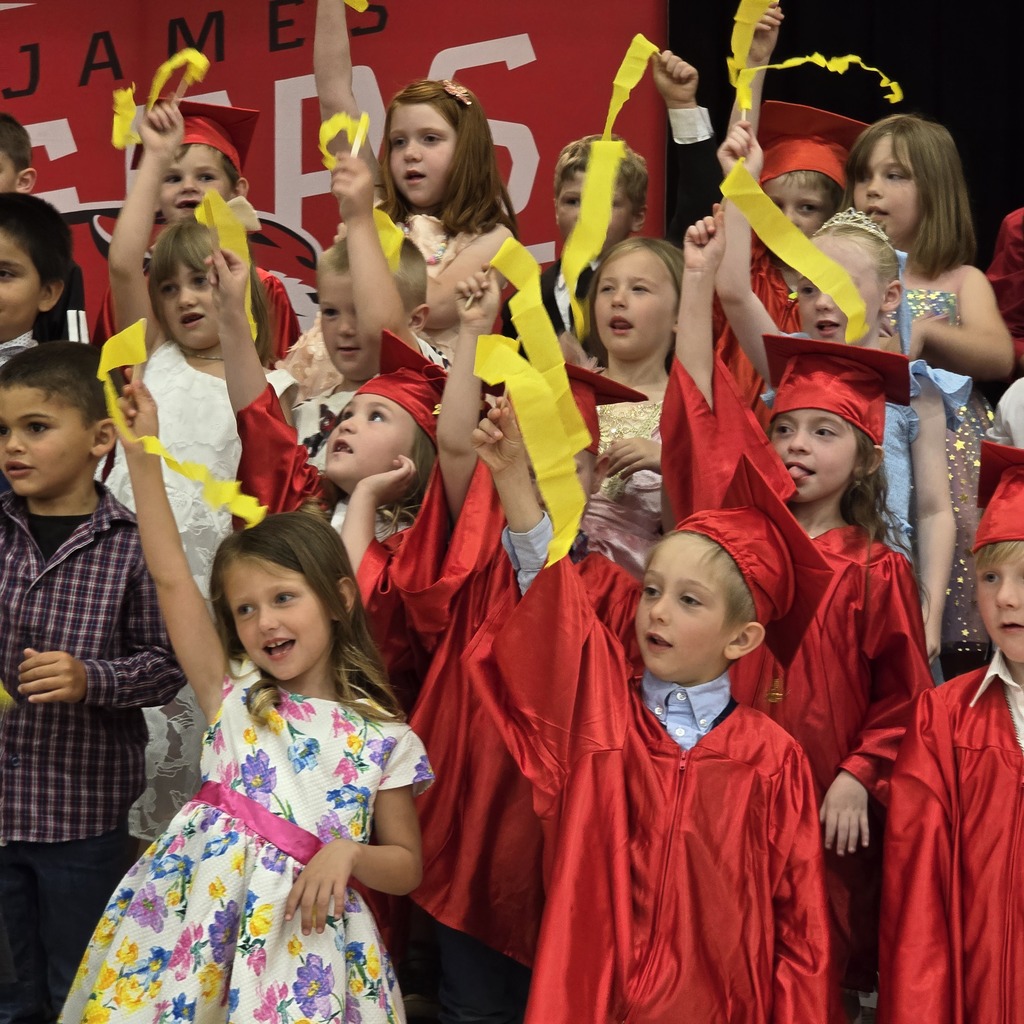 Children in red graduation gowns and caps at an event. Some hold yellow ribbons. A red banner with white text is visible in the background.
