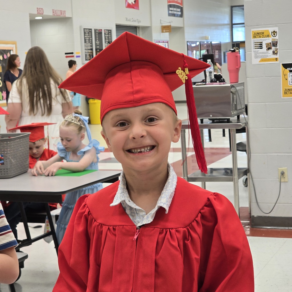 A young boy in a red graduation gown and cap smiles at the camera. Behind him, two children sit at a table.